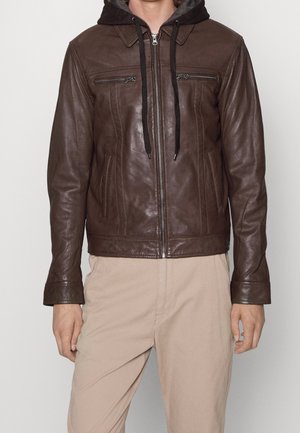 Man draagt een donkerbruine leren jas met rits met een zwarte hoodie eronder en beige broek, staand tegen een neutrale achtergrond.