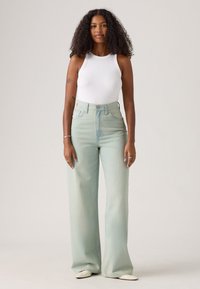 Light blue high-waisted jeans made of denim, featuring a wide leg design. Paired with a ribbed white tank top and cream-colored shoes.