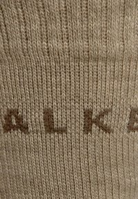 Beige gebreide sok met een geribbelde structuur en lichtbruine letters. Het materiaal lijkt zacht en elastisch met een gelijkmatig stekenpatroon.