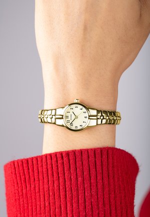 Montre-bracelet dorée avec un cadran rond, un fond blanc, des index horaires noirs et un bracelet texturé, portée sur un poignet nu.