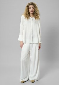 Weißes Seidenhemd mit Knopfleiste und langen Ärmeln, kombiniert mit lockeren, weit geschnittenen weißen Hosen. Sanfte Textur, minimalistisches Design und leichtes Material.