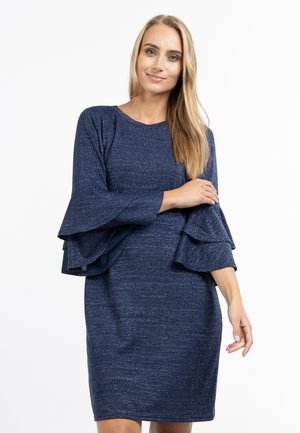 Kobieta z długimi blond włosami ubrana w granatową sukienkę z warstwowymi rozszerzanymi rękawami, stojąca na białym tle.