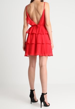 Femme portant une robe rouge à volants avec un dos en V profond et des fines bretelles, associée à des chaussures noires à talons hauts avec bride à la cheville.