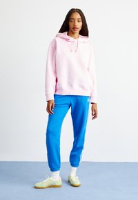 Ροζ φούτερ με κορδόνια, χαλαρή εφαρμογή. Με μπλε φόρμες με λευκές λωρίδες στο πλάι. Ανοιχτό πράσινα παπούτσια, υφασμάτινη υφή, casual σχεδίαση.