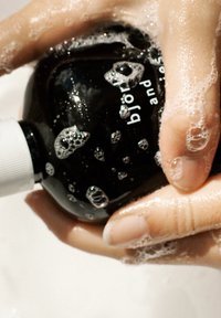 Schwarze Flasche mit einem weißen Pumpspender, von Blasen bedeckt, in Händen gehalten. Etikett teilweise sichtbar, mit dem weißen Text "björk".