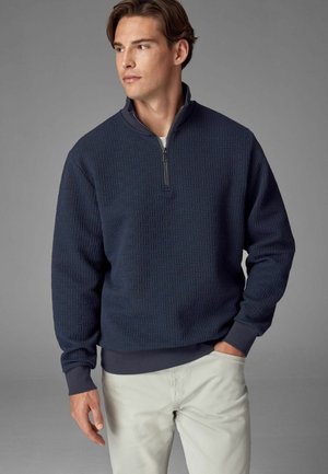 Navy pullover sweatshirt with a textured woven pattern, half-zip collar, and ribbed cuffs and hem. Paired with light-colored pants.