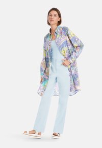 Mehrfarbiges gemustertes Hemd mit langen Ärmeln und lockerer Passform, kombiniert mit hellblauen hoch taillierten Hosen und weißen Sandalen.