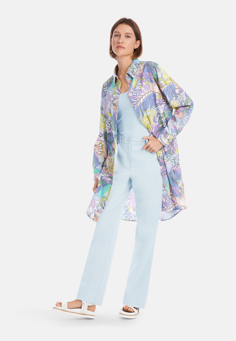 Mehrfarbiges gemustertes Hemd mit langen Ärmeln und lockerer Passform, kombiniert mit hellblauen hoch taillierten Hosen und weißen Sandalen.