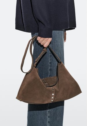 Person holding a brown suede handbag with metal studs, wearing a navy sweater and blue denim jeans against a plain background.