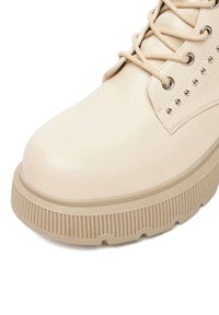 Beżowy botek z ekoskóry sięgający kostki, z okrągłym noskiem, teksturowaną podeszwą platformową, sznurowanym zapięciem i dekoracyjnymi ćwiekami z boku.