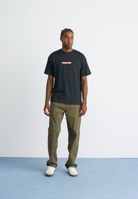 Man wearing black graphic t-shirt, olive cargo pants, and white sneakers standing on blue floor against white wall, looking slightly right.