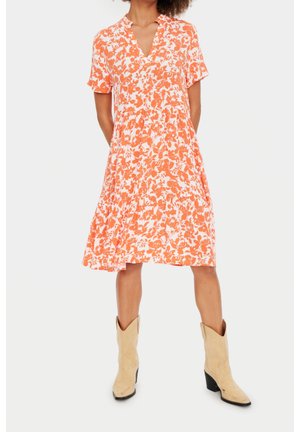 Femme portant une robe courte à manches courtes à fleurs orange et blanche, associée à des bottes de cowboy beiges mi-mollet, debout, les mains dans le dos.