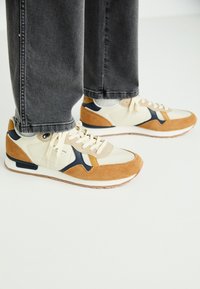 Baskets beiges et brunes en suède et maille, avec des accents bleu marine et beige, à bout rond, semelle blanche et design à lacets.