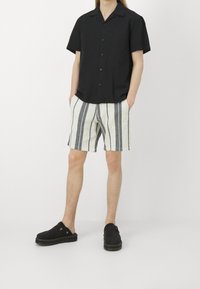 Person klädd i en svart kortärmad skjorta med knappar, vita shorts med vertikala svarta ränder och svarta slip-on skor mot vit bakgrund.