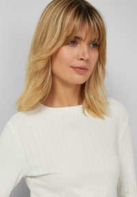 Weißes, geripptes Langarmshirt mit rundem Ausschnitt. Haare sind in sanften Wellen mit geraden Ponyfransen gestylt. Weiche Textur und körpernahe Passform.