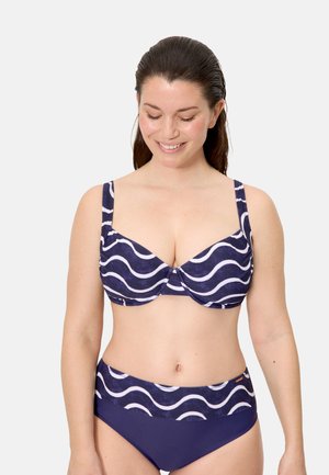 Mujer con un bikini azul marino con rayas onduladas blancas en la parte superior y la cintura, sonriendo con los ojos cerrados frente a un fondo claro.