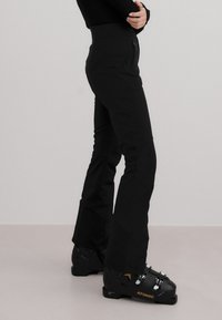 Person iført svarte skibukser og svarte Atomic skistøvler, stående mot en ensfarget lys grå bakgrunn.