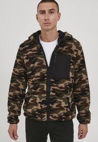 Fleece jacket in camouflage pattern with black zippered chest pocket. Features a zip closure and a hood, showcasing a soft, textured fabric.