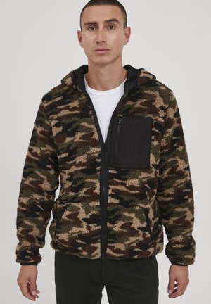 Fleece jacket in camouflage pattern with black zippered chest pocket. Features a zip closure and a hood, showcasing a soft, textured fabric.