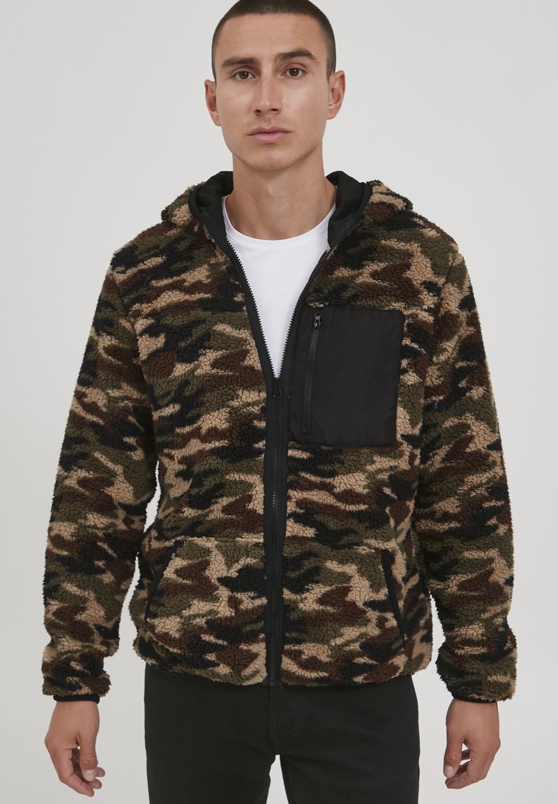 Fleece jacket in camouflage pattern with black zippered chest pocket. Features a zip closure and a hood, showcasing a soft, textured fabric.