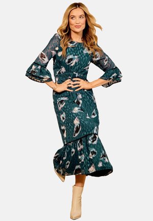 Robe à motif teal avec des manches évasées, un ourlet en volants, des motifs à pois et floraux, associée à des bottines beige à la cheville.