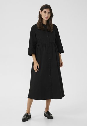 Black mid-length dress in a loose fit with a button-down front and three-quarter sleeves, made of smooth fabric. Paired with black loafers.