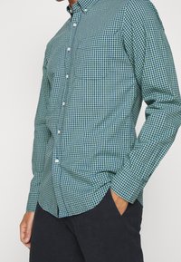 Grün-blau kariertes Hemd mit Knöpfen, tailliert, langärmlig, mit einer Brusttasche und weißen Knöpfen. Schlichte schwarze Hose.