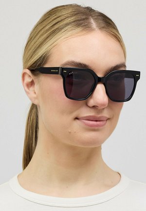 Frau mit nach hinten gebundenen blonden Haaren, die große schwarze quadratische Sonnenbrille und ein weißes Oberteil vor neutralem Hintergrund trägt.