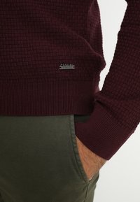 Homme portant un pull texturé bordeaux avec une étiquette "Casual Friday" et un pantalon vert olive, main reposant dans la poche.