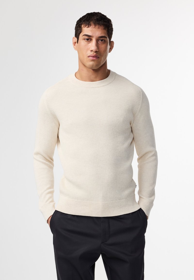 Hellbeige, gerippter Strickpullover mit rundem Halsausschnitt und langen Ärmeln, kombiniert mit dunklen Hosen. Der Stoff hat eine weiche Textur.