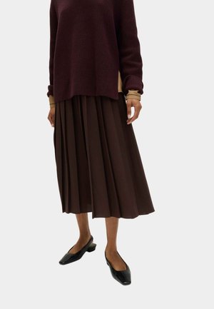 Bordeauxroter Strickpullover mit langen Ärmeln, darüber getragen ein brauner plissierter Rock. Das Outfit wird mit schwarzen spitzen Ballerinas kombiniert.