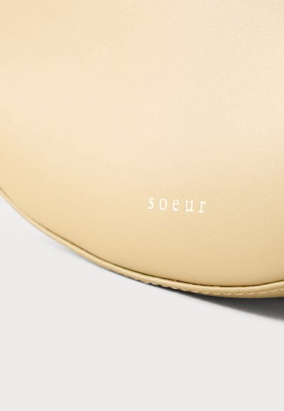 Μπεζ στρογγυλεμένο δερμάτινο αντικείμενο με λευκή μικρή πεζή γραφή "soeur" ανάγλυφη κοντά στην άκρη σε απλό ανοιχτό φόντο.