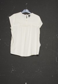Blusa corta a maniche corte color crema con pizzo sui maniche e sul collo, appesa a una gruccia trasparente su uno sfondo nero testurizzato.