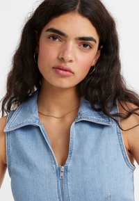 Colete de jeans azul claro com fecho éclair à frente, grande colarinho e sem mangas. Costura visível e textura suave. Corrente dourada visível no pescoço.