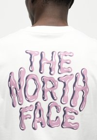 Οπίσθια όψη ατόμου που φορά λευκό πουκάμισο με μεγάλο ροζ, γυαλιστερό, κείμενο σε στυλ λεκέ που γράφει "THE NORTH FACE" στην πλάτη.