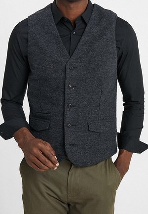 Mand iført mørkegrå knappet vest over sort skjorte og olivengrønne bukser, med den ene hånd i lommen og den anden nær taljen.