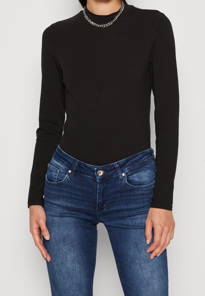 Schwarzes Langarmoberteil aus weichem Stoff, kombiniert mit dunkelblauen Jeans. Das Oberteil hat einen hohen Kragen und eine schlanke Passform.