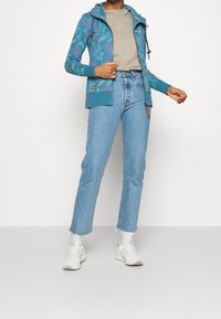 Sudadera con cremallera en tono azul verdoso y patrón floral, combinada con jeans rectos de color azul claro. Lleva zapatillas blancas y una camiseta beige por debajo.