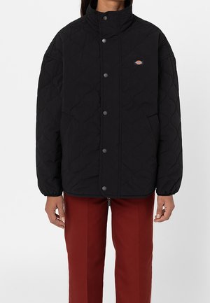 Personne portant une veste matelassée noire avec des boutons-pression et un petit logo sur la poitrine, associée à un pantalon droit rouge.