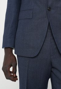 Costum bleumarin dintr-un material texturat, sacou cu un singur rând de nasturi și rever ascuțit, și pantaloni ajustați cu buzunare laterale.
