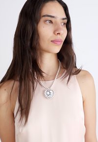 Silver heart-shaped necklace with a large crystal centerpiece, surrounded by small beads, worn over a light pink, sleeveless blouse.