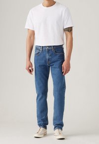 Siniset farkut suoralla leikkauksella, klassisella viiden taskun mallilla ja yksinäppäimellä suljettuna, yhdistettynä yksiväriseen valkoiseen pyöreäkaulaiseen t-paitaan.