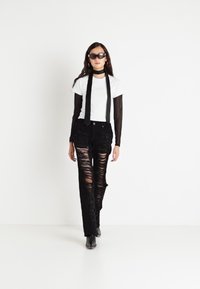 Pantalones vaqueros negros rasgados de cintura alta, combinados con una camiseta blanca con logo y una blusa de manga larga de malla negra. La modelo lleva gafas de sol y botines.