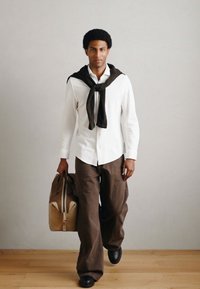 Valge puuvillane särk, kaela ümber seotud pruun kampsun, pruunid laia säärega püksid ja mustad kingad, kaasas beež dufflikott.