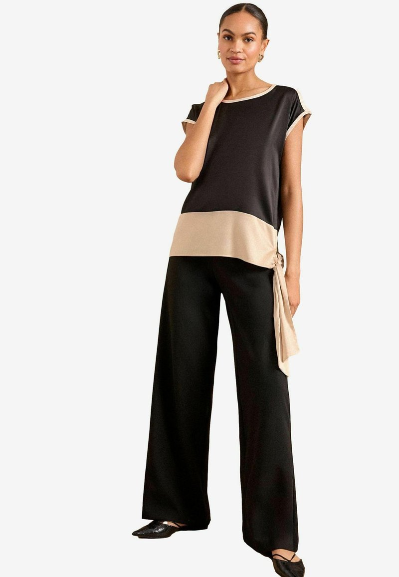 Blusa nera e beige con maniche corte, caratterizzata da un dettaglio con nodo laterale. Abbinata a pantaloni neri a gamba larga e scarpe nere basse.