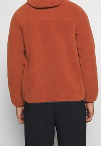 Giacca in pile color ruggine con cappuccio, caratterizzata da una texture morbida, polsini elasticizzati e un orlo dritto. Progettata per il comfort e il calore.