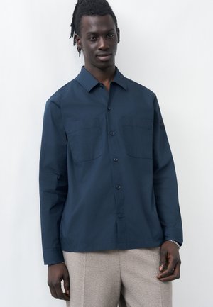 Νεαρός άνδρας με σκουρόχρωμο δέρμα και κοντά dreadlocks, φορώντας σκοτεινό μπλε πουκάμισο με κουμπιά και ανοιχτό μπεζ παντελόνι, στέκεται μπροστά σε έναν απλό λευκό φόντο.