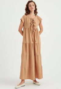 Maxikleid in Hellbraun, aus strukturiertem Stoff mit gebundenem Ausschnitt und kurzen Ärmeln. Hat ein gestuftes Design und reicht bis zum Boden.