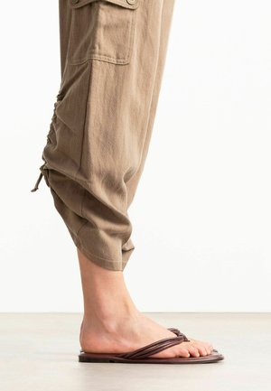 Bloot voet met een bruine platte sandaal met elastische riempjes, gecombineerd met beige taps toelopende broek, opgerold bij de enkel.