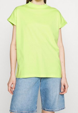 Personne portant un haut ample à manches courtes vert citron et un short en jean bleu clair arrivant aux genoux, debout devant un fond uni.